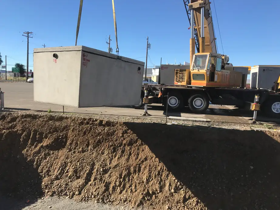 A precast concrete septic tank on the boom of a yellow crane truck beside the excavation