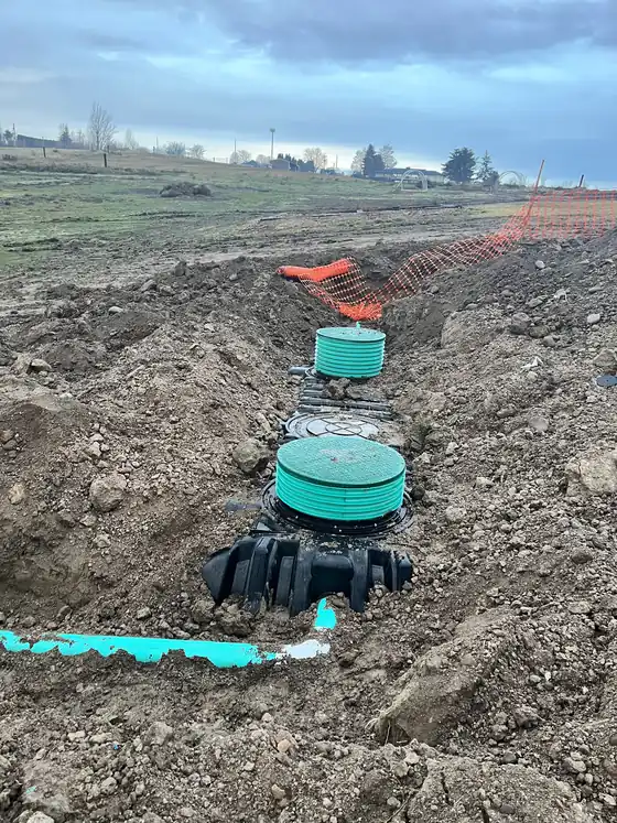 Green-lidded tank risers standing in an open excavation behind orange safety fence, with flat fields beyond