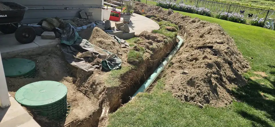 A trench cut across a backyard with a green riser lid and teal pipe laid in it beside the corner of a house
