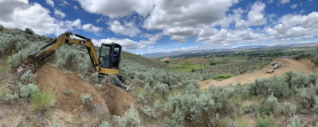 A yellow compact excavator digging a test pit on an open sagebrush hillside above the valley